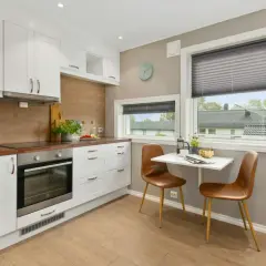 Moderna cocina equipada con gabinetes blancos, encimeras de madera y un horno. Una pequeña mesa con sillas marrones está junto a la ventana.