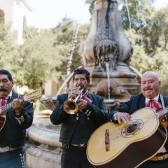 Trzech muzyków mariachi gra na gitarach i trąbce przy fontannie.