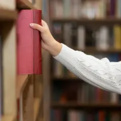 Ręka sięga po czerwoną, grubą książkę. Może to być ciekawa książka dla starszej osoby, pełna wspomnień lub historii.