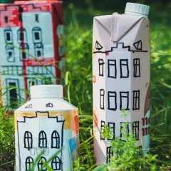 Opakowania po mleku w kształcie domków stoją w trawie. Zastanawiasz się, gdzie wyrzucać opakowanie po mleku? Do plastiku lub papieru, w zależności od oznaczeń.