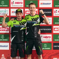 Dwóch zawodników Energylandia Rally Team pozuje z podniesionymi pięściami na tle baneru z logo sponsorów.