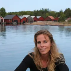 Woman with long blonde hair smiling, with red boathouses on a rocky shore in the background.