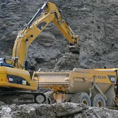 Żółta koparka CAT zasypuje ziemią wywrotkę Volvo. Zastanawiasz się, ile kosztuje godzina pracy koparki?