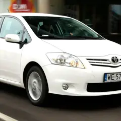 Biała Toyota Auris w ruchu miejskim, z widocznym kierowcą za kierownicą.
