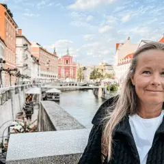 Kvinna ler vid floden i Ljubljana, Sloveniens huvudstad, med färgglada byggnader och en kyrka i bakgrunden.