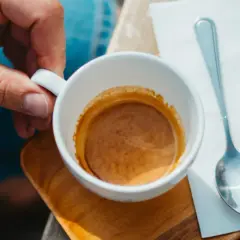 Dłoń trzyma filiżankę z espresso. Idealna ilość kawy na espresso to około 7-9 gramów. Obok leży łyżeczka i szklanka wody.