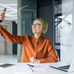 Uśmiechnięta blondynka w okularach robi sobie zdjęcie telefonem przy biurku.