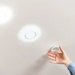 Mano instalando una lámpara techo redonda. Dos luces circulares ya iluminan el techo blanco.