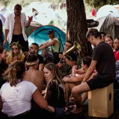 Grupa ludzi relaksuje się na festiwalu Pannonica, jeden z nich gra na puzonie. W tle widać namioty.