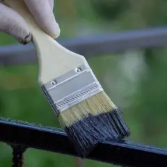 Mano guantata che usa un pennello per verniciare una ringhiera di metallo scuro.