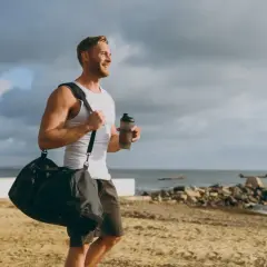 Uśmiechnięty mężczyzna z torbą sportową i bidonem na plaży. Zastanawia się, glutamina co to, czy pomoże mu w regeneracji po treningu.