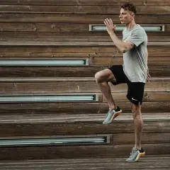 Mężczyzna w szarej koszulce i czarnych spodenkach wykonuje dynamiczne ćwiczenie, podnosząc kolano. Intensywny trening HIIT na drewnianych schodach.