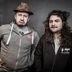 Dwóch mężczyzn, jeden w kapeluszu i z miną figo, drugi z burzą loków, pozuje do zdjęcia.