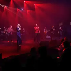 Zespół muzyczny na scenie, oświetlony czerwonymi światłami. Wokalistka w granatowej sukni, obok niej gitarzyści i wokalista w czerwonej koszuli. W tle widać perkusję i innych muzyków. Publiczność siedzi w ciemności. To jakby **sufler** podpowiadał im k...
