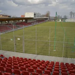 Puste trybuny i zielona murawa na stadionie w Niepołomicach. W tle budynek klubowy i balon nad boiskiem.