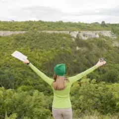 Kobieta z mapą i kubkiem w rękach, z ramionami rozłożonymi na tle zielonych wzgórz. To kwintesencja ekoturystyki – co to znaczy? Cieszyć się naturą!