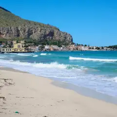 Spiaggia dorata e mare turchese in Sicilia, con un borgo colorato incastonato tra la montagna e l'acqua cristallina.