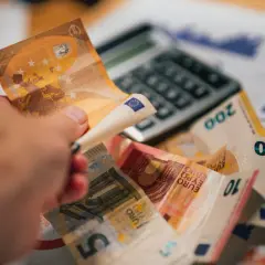 Dłoń trzyma banknot 50 euro nad stosem innych banknotów i kalkulatorem. To symbol prowizji i finansów.