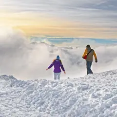 Dwie osoby wędrują po ośnieżonym szczycie. W oddali widać góry i chmury. Czyżby szukali odpowiedzi na pytanie: jakie raki w Tatry?