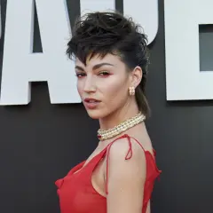 Mujer con un atrevido corte mulet y maquillaje dramático, luciendo un vestido rojo y joyas doradas.