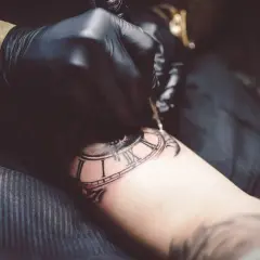 Artysta w czarnej rękawiczce tworzy tatuaż zegara, używając techniki whip shading do dodania głębi i cieni.