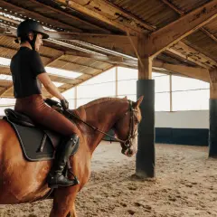 Kobieta w kasku i brązowych bryczesach dosiada kasztanowego konia w ujeżdżalni. To część jej kursu instruktora jazdy konnej.