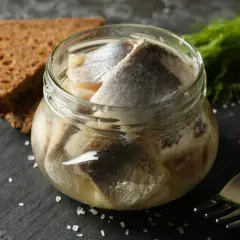 Słoik śledzi w oleju, jak u babci. Obok kromki chleba i widelca.