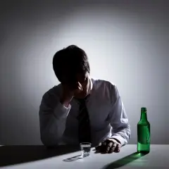 Zmartwiony mężczyzna w białej koszuli i krawacie, z ręką na czole, siedzi przy stole z butelką zielonego alkoholu i kieliszkiem. Zastanawia się, ile trwa zatrucie alkoholowe.