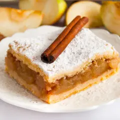 Kawałek babcinej szarlotki najlepszej, posypanej cukrem pudrem i ozdobionej laskami cynamonu, na talerzyku z jabłkami w tle.