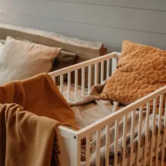 Ett vitt spjälsängsskydd med mjuka filtar och kuddar i varma färger. Perfekt för en mysig barnkammare.