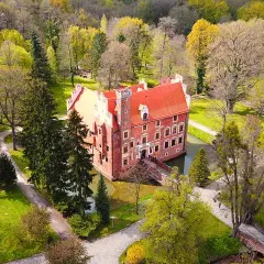 Zamek na wodzie w Wojnowicach otoczony wiosenną zielenią i drzewami. Czerwona bryła zamku z charakterystycznym dachem odbija się w spokojnej tafli wody.