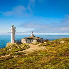 Faro blanco y edificio antiguo en Cabo Touriñán, con mar azul y cielo nublado.