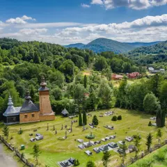 Drewniana cerkiew i cmentarz w malowniczej dolinie Beskidu Niskiego. W oddali widać zalesione wzgórza i domy.