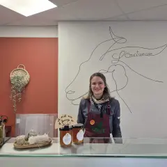 Une vendeuse souriante derrière un comptoir, avec des cornichons, du miel et du fromage de chèvre, peut-être du fromage Saint-Aubin, en vente.