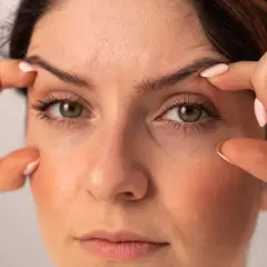 Une femme examine son visage, ses doigts effleurant sa peau. Elle semble préoccupée car j'ai des cernes noires sous les yeux.