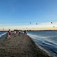 Ludzie spacerują po piaszczystej mierzei, podczas gdy na niebie widać liczne latawce do kitesurfingu.