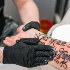 Ręce w czarnych rękawiczkach smarują świeży tatuaż na przedramieniu. Pielęgnacja jest kluczowa, by dowiedzieć się, jak długo smarować tatuaż.