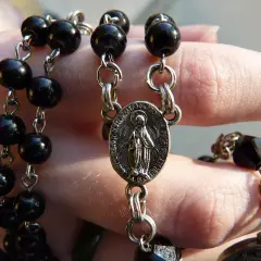 Dłoń trzyma czarny różaniec z medalikiem Matki Boskiej. Czas modlitwy, ile trwa różaniec na pogrzebie, zależy od intencji.