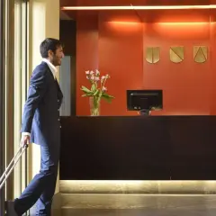 Uomo con valigia rossa alla reception, pronto per l'early check-in. La receptionist sorride accogliendolo.