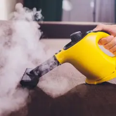 Mano sujetando una limpiadora a vapor amarilla, desprendiendo vapor sobre un sofá marrón.