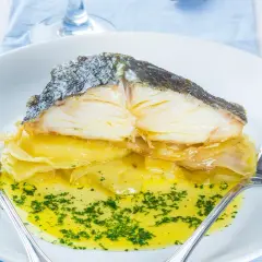 Merluza al horno con patatas, jugosa y dorada, servida sobre una cama de patatas y cebolla caramelizada, bañada en aceite de oliva y perejil picado.