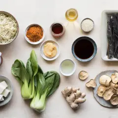 A collection of ingredients for a Japanese meal, including noodles, bok choy, tofu, ginger, dried mushrooms, and various Japanese sauces and seasonings.