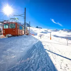 Bernina Express-tåget står parkerat i snön, redo att ta passagerare genom ett snöigt alpint landskap under en klarblå himmel.