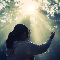 Una niña con coleta, con las manos alzadas, parece soñar con espíritus mientras la luz del sol atraviesa los árboles.