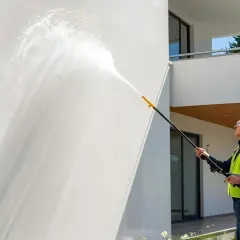 Mężczyzna w kamizelce odblaskowej myje białą elewację domu myjką ciśnieniową.