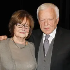 Anna Opani w okularach i z perłami na szyi, obok elegancko ubranego mężczyzny w garniturze.