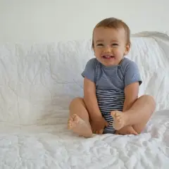 Lächelndes Baby im gestreiften Body sitzt auf weißer Decke.