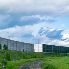 Długie ekrany akustyczne drogowe chronią przed hałasem. Betonowe i zielone panele wzdłuż drogi, na tle błękitnego nieba z chmurami.