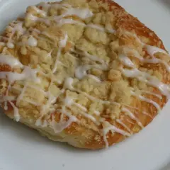 Szneka z glancem, puszyste ciasto drożdżowe z kruszonką i lukrem, wygląda apetycznie na białym talerzu.