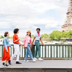 Quatre jeunes amis profitent d'une promenade ensoleillée à Paris, avec la Tour Eiffel en arrière-plan. L'un d'eux, un étudiant, porte un foulard rose vif.